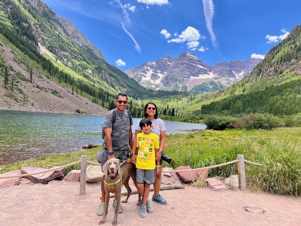 Riddhi, Srini and their son smiling on a Colorado mountain trail with their grey dog, pine trees and a green valley behind them.