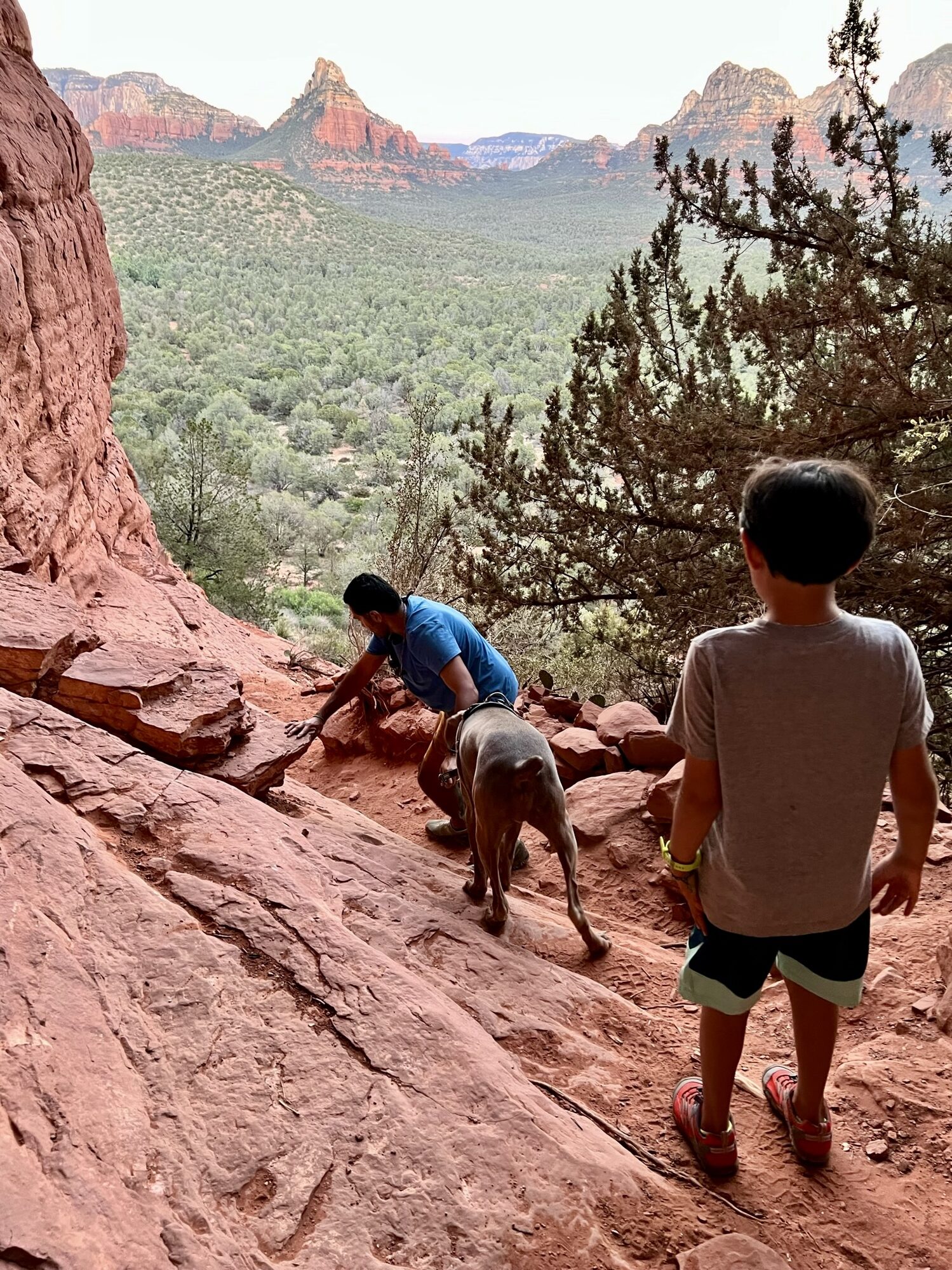 Man and dog scrambling up red rock toward the Birthing Cave, with a boy following and red buttes beyond