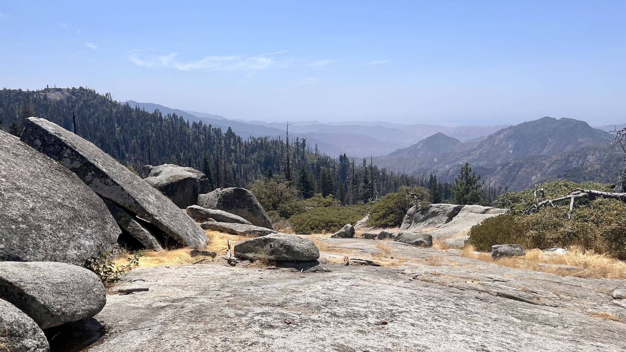 Large granite boulders on a wide rock slab at Beetle Rock, overlooking forested foothills and distant hazy ridges.