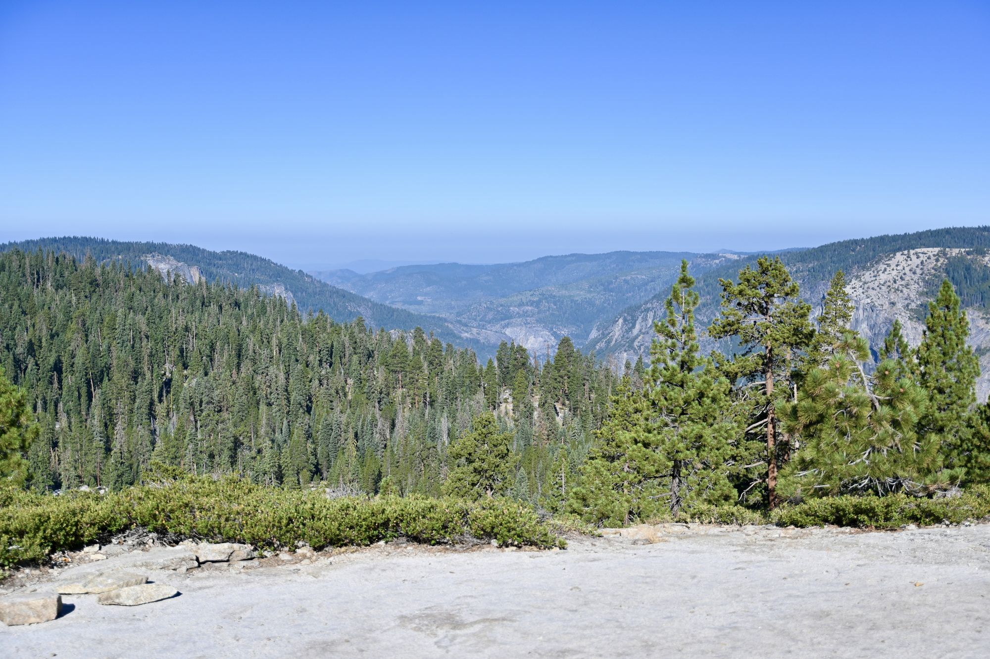 A wide granite slope in the foreground, with pine forest and layered blue mountain ridges beyond it.
