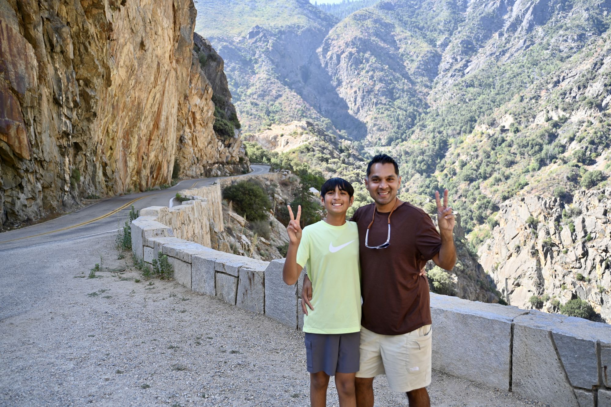Our son and his dad by a stone wall at a roadside pullout, with a cliff-lined road curving into the canyon behind them.
