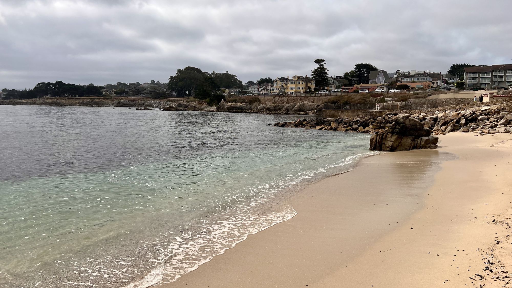 A small sandy beach with clear, shallow water and a rocky point, with houses and cypress trees beyond.