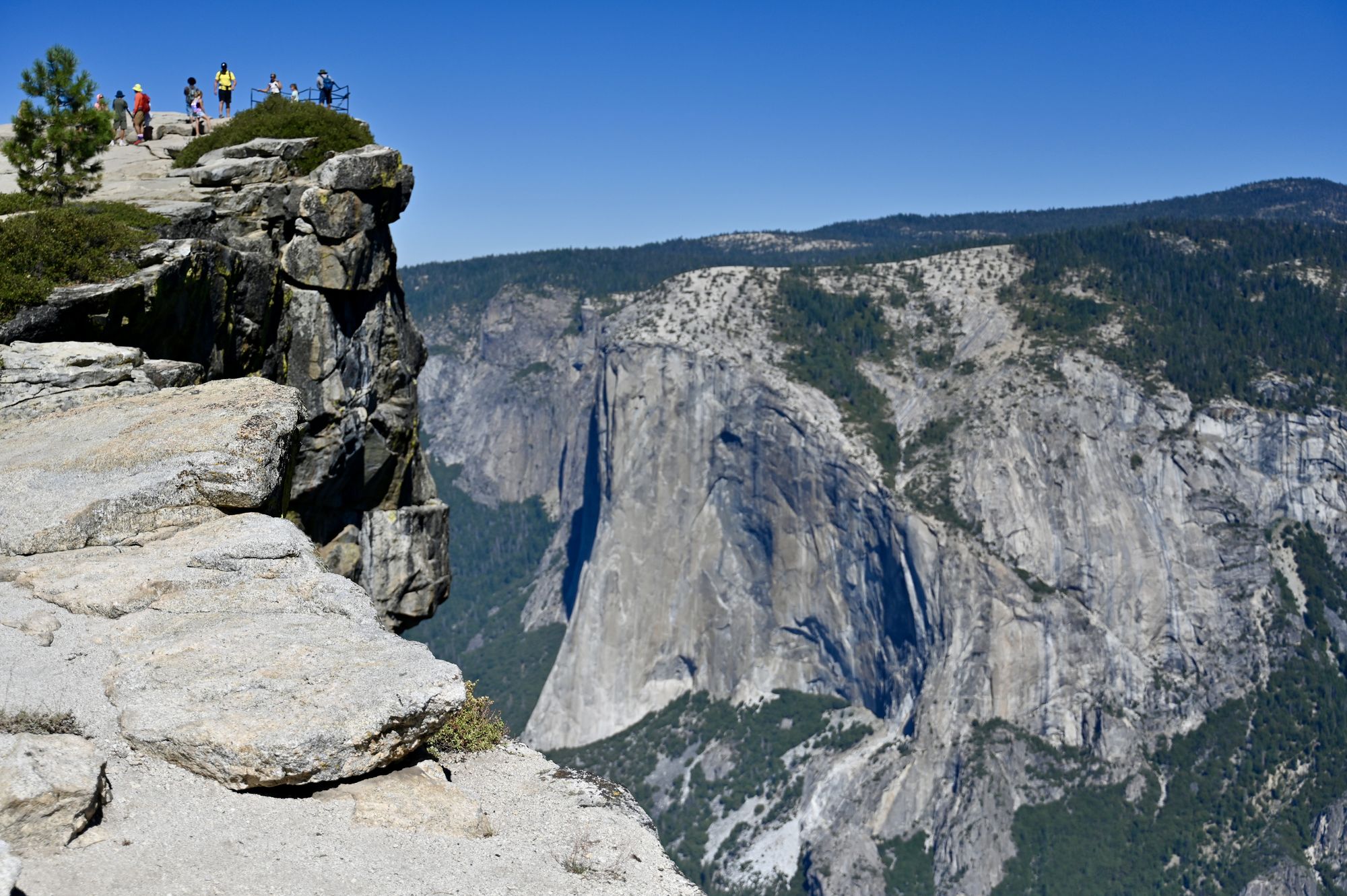 A rocky outcrop with a railing and a small group of people at the edge, looking across a deep valley at El Capitan.