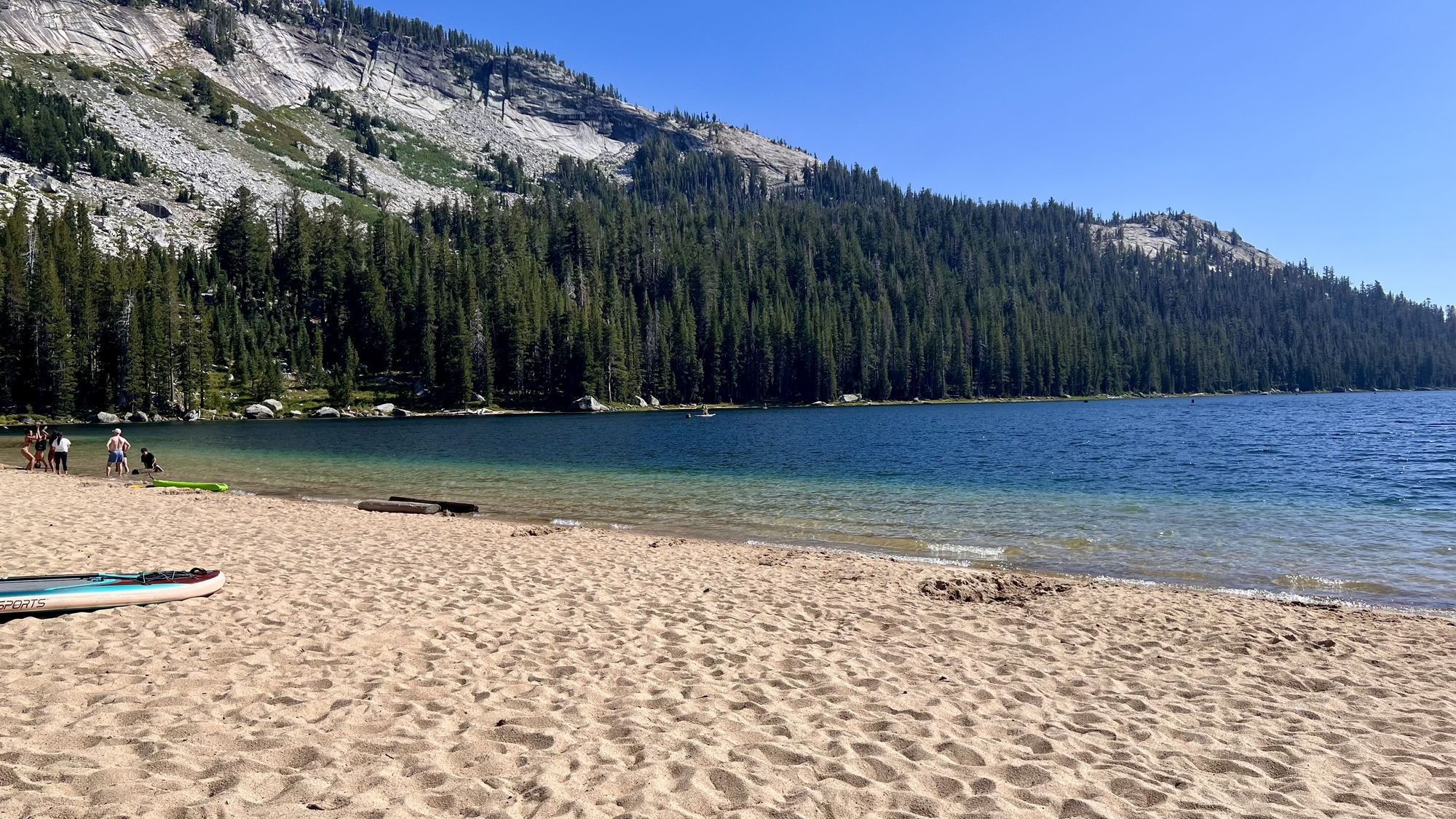 The sandy beach at Tenaya Lake, with a paddleboard on the sand, a few families at the water's edge and a granite ridge above the trees.