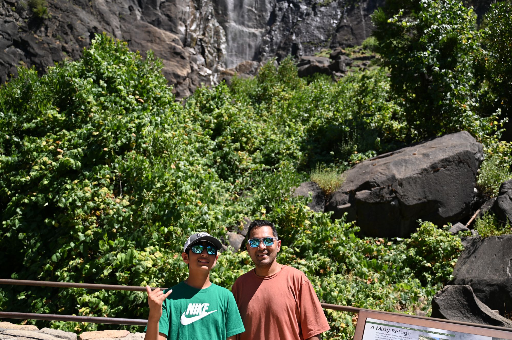 Our son and his dad at the Bridalveil falls viewpoint railing, with dense green shrubs and a dark granite wall behind them.