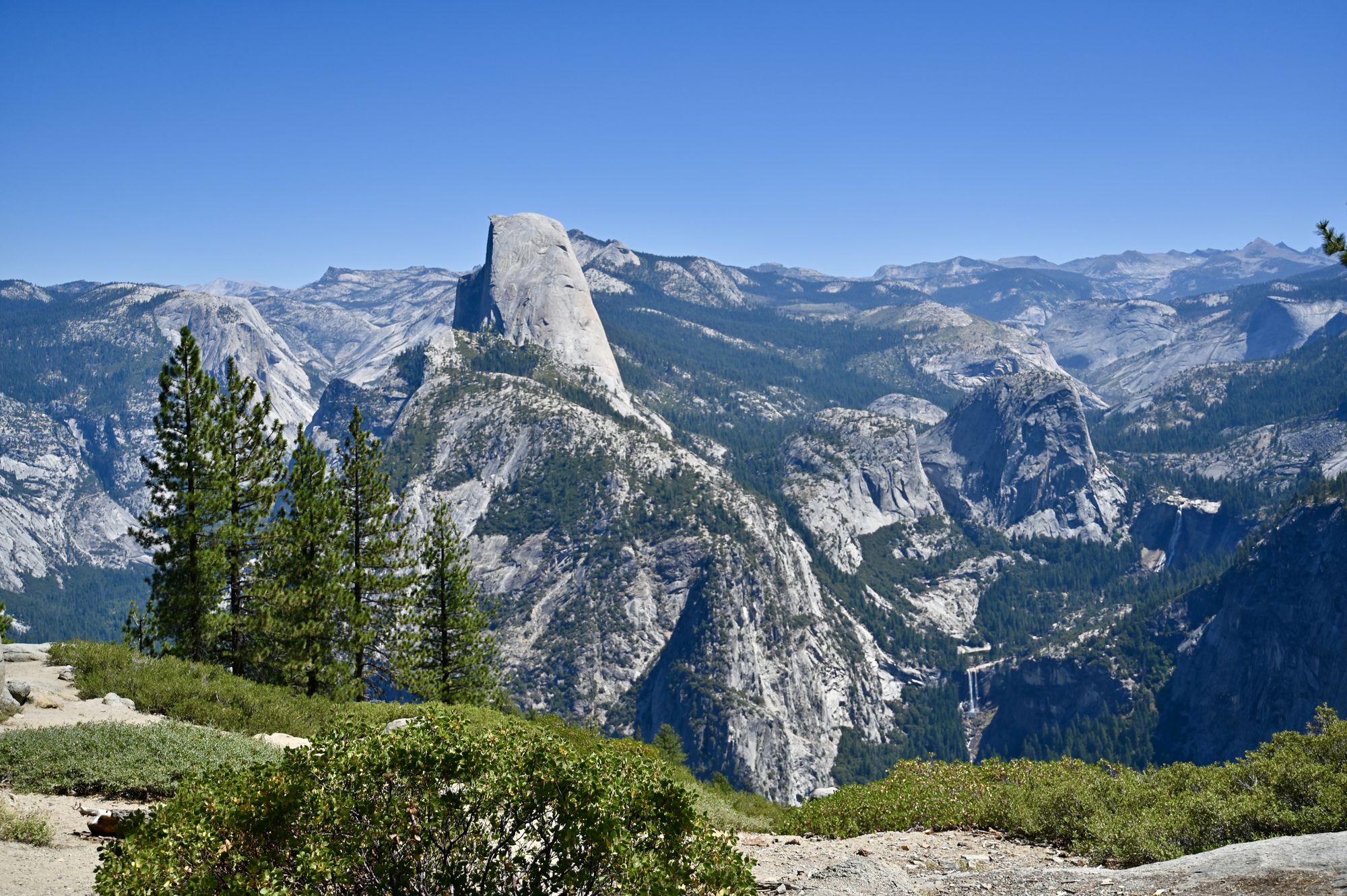 Half Dome rising above a forested granite valley, with Vernal falls and Nevada falls visible as thin white ribbons on the right.