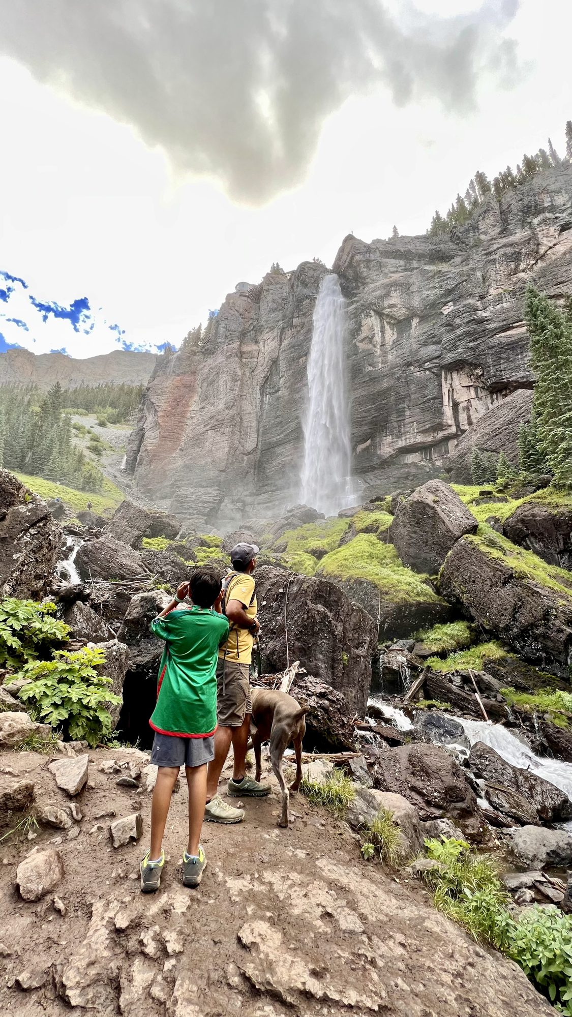 Boy and dog on a rocky trail looking up at Bridal Veil Falls pouring off a tall cliff