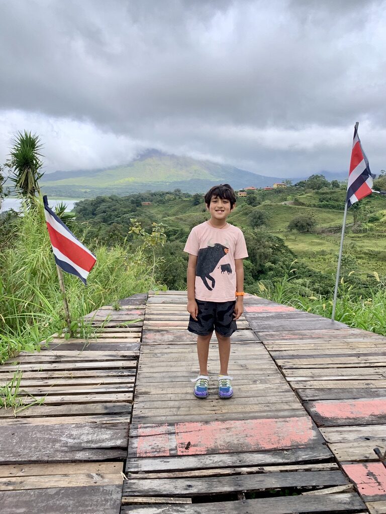 Boy standing on a wooden deck between two Costa Rican flags, with the lake and volcano behind