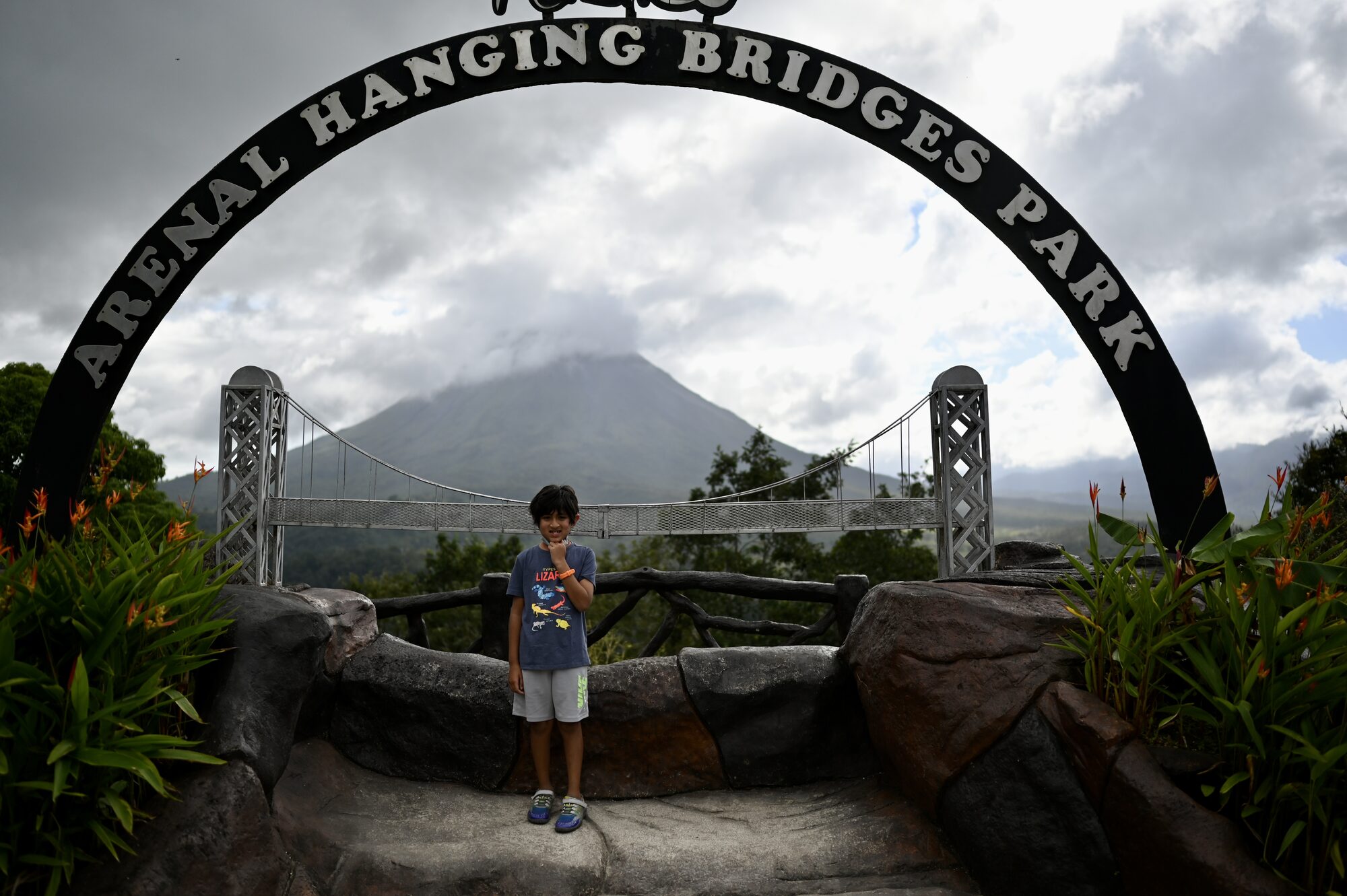 Boy standing under the Arenal Hanging Bridges Park arch, with the volcano in cloud behind