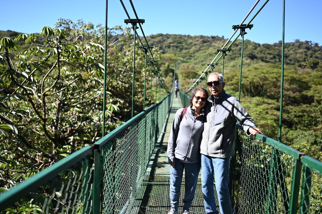 An older couple smiling on a suspension bridge above the treetops