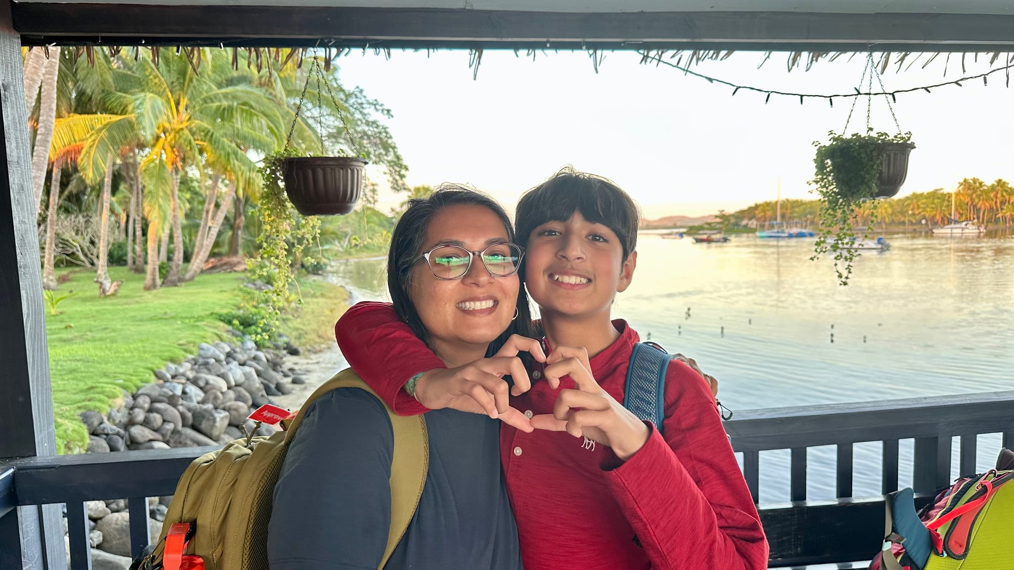 Our son and I making a heart with our hands on a covered jetty, backpacks on, with a calm lagoon, moored boats and palm trees behind us in the early-morning light.