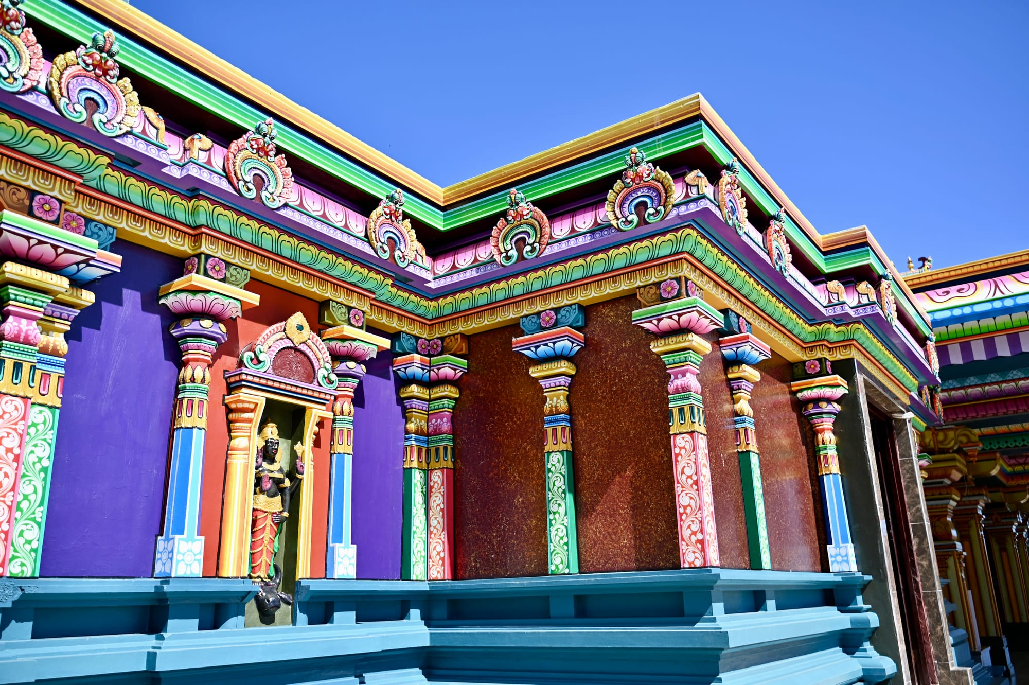 The outer wall of the Sri Siva Subramaniya temple, painted purple, red, green and gold, with ornate carved pillars, a deity statue in an alcove and decorative crests along the roofline against a clear blue sky.