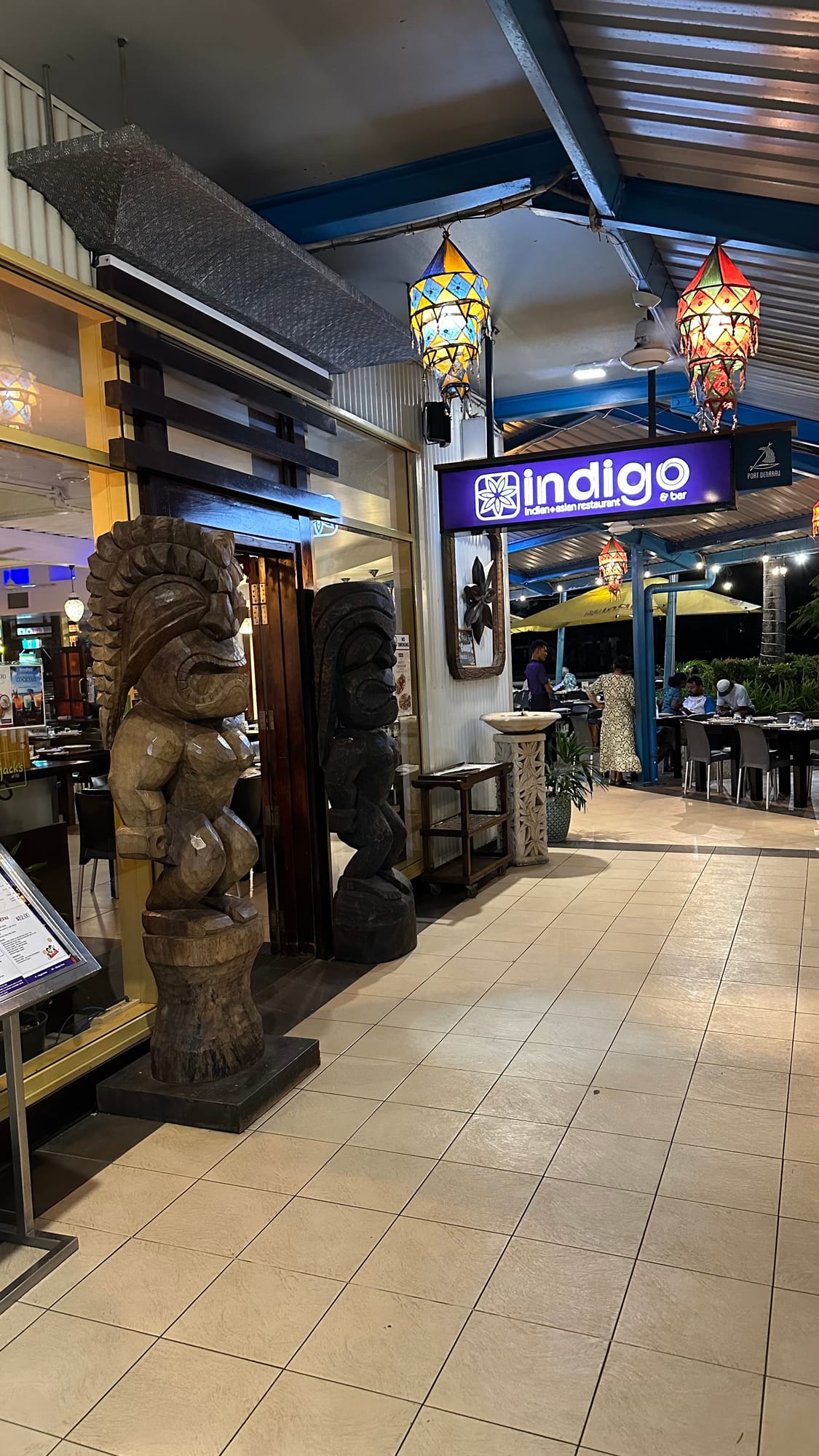 The entrance to Indigo restaurant along a tiled walkway, flanked by tall carved wooden tiki figures, with coloured glass lanterns hanging above and the lit dining room beyond.