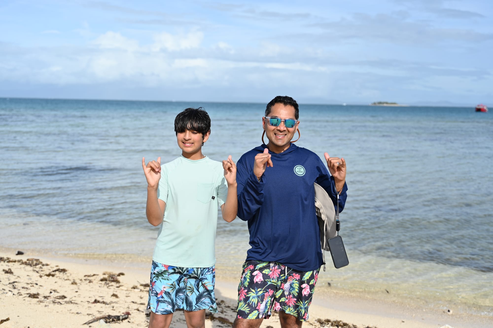 Our son and his dad standing on the beach in matching floral swim shorts, throwing shaka signs, with calm blue water and a small island on the horizon.