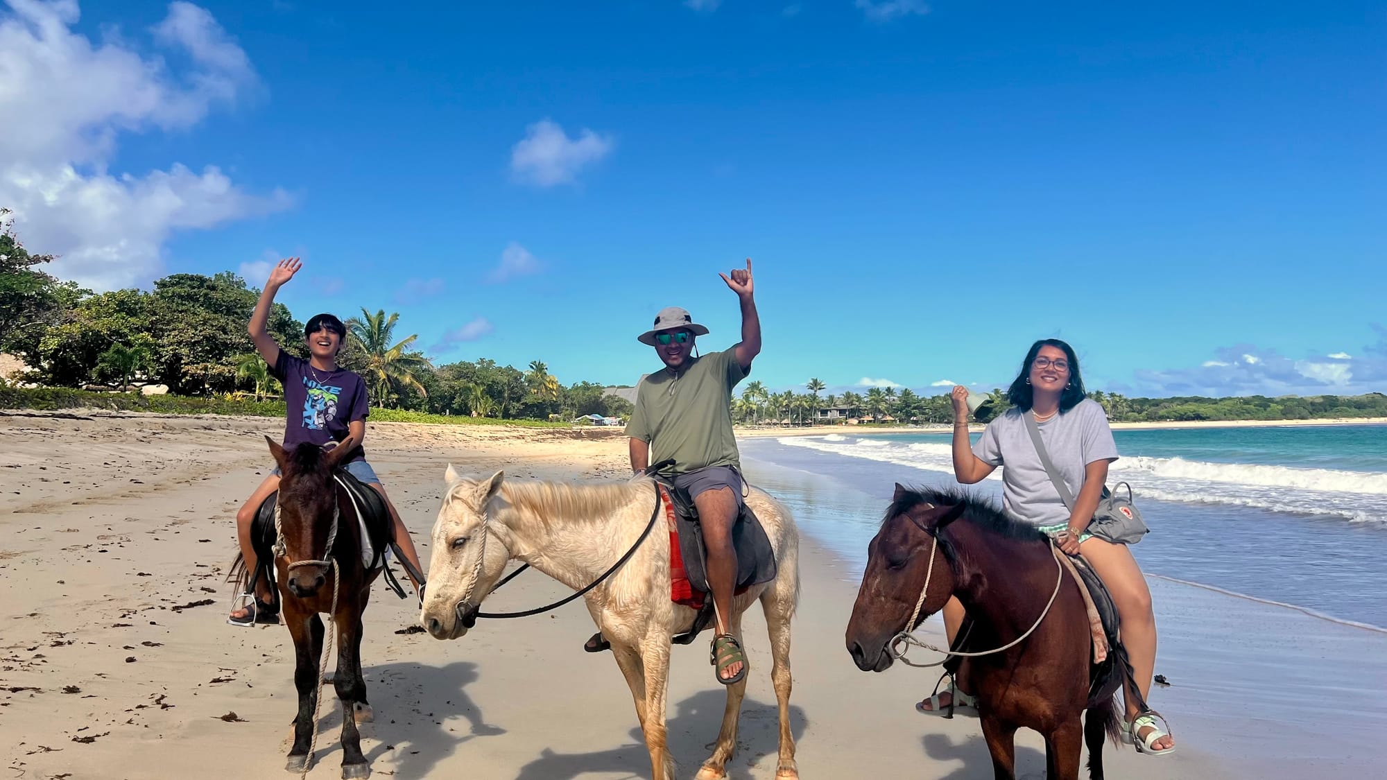Our son, his dad and I on three horses at the water’s edge, all waving, with the beach and palm trees stretching behind us.