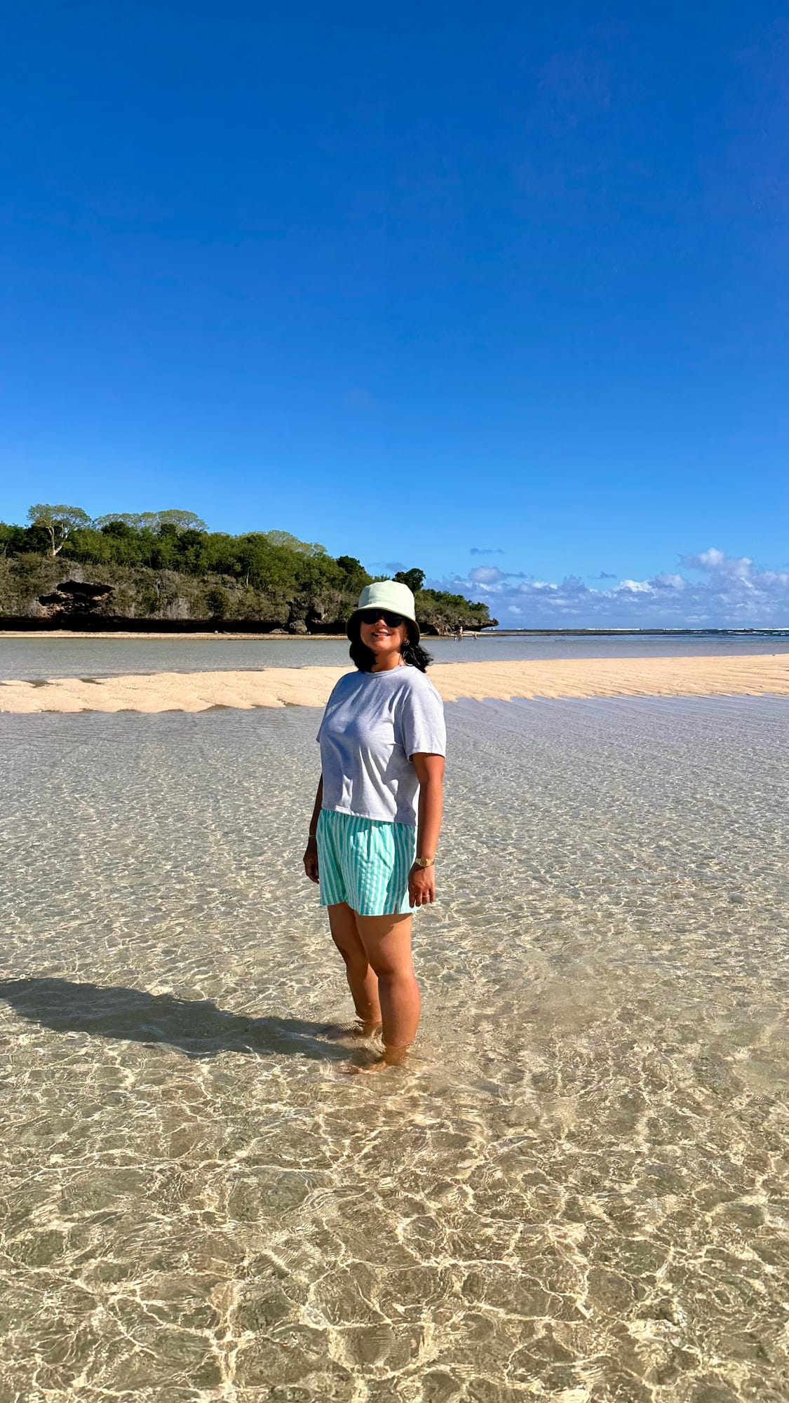 Me standing ankle-deep in clear, rippled shallow water in a sun hat, with a sandbar and a small, tree-covered island behind.