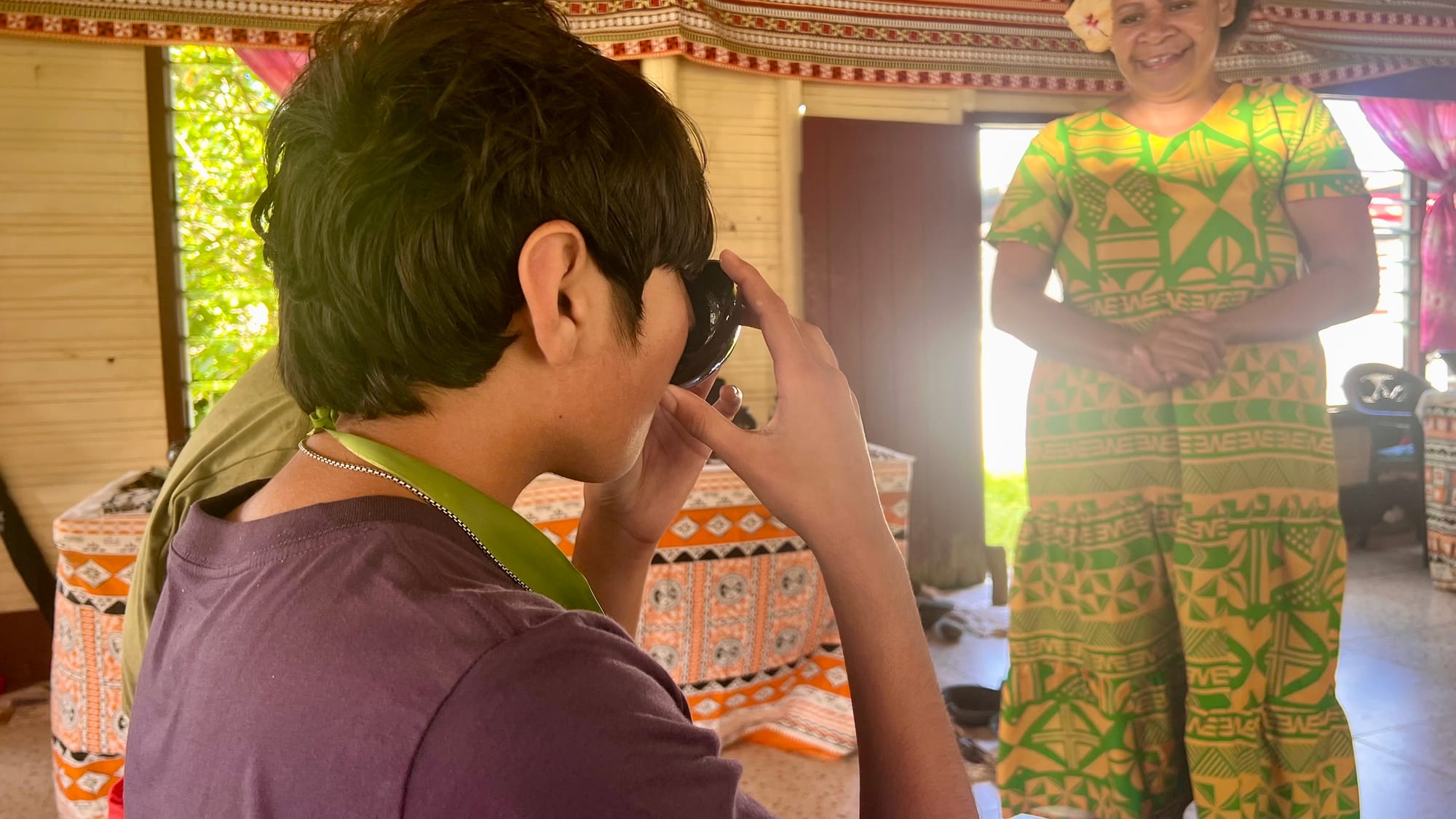 Our son raising a small bowl to drink during the kava ceremony, as a smiling village woman in a bright green and yellow patterned dress looks on.