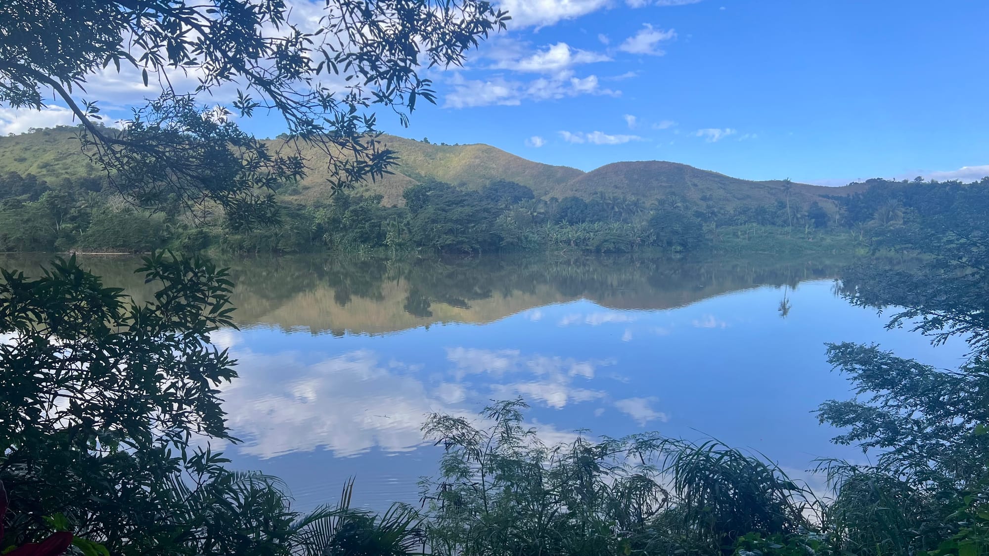 A wide, still river mirroring the green hills and blue sky, seen through overhanging branches from the village bank.