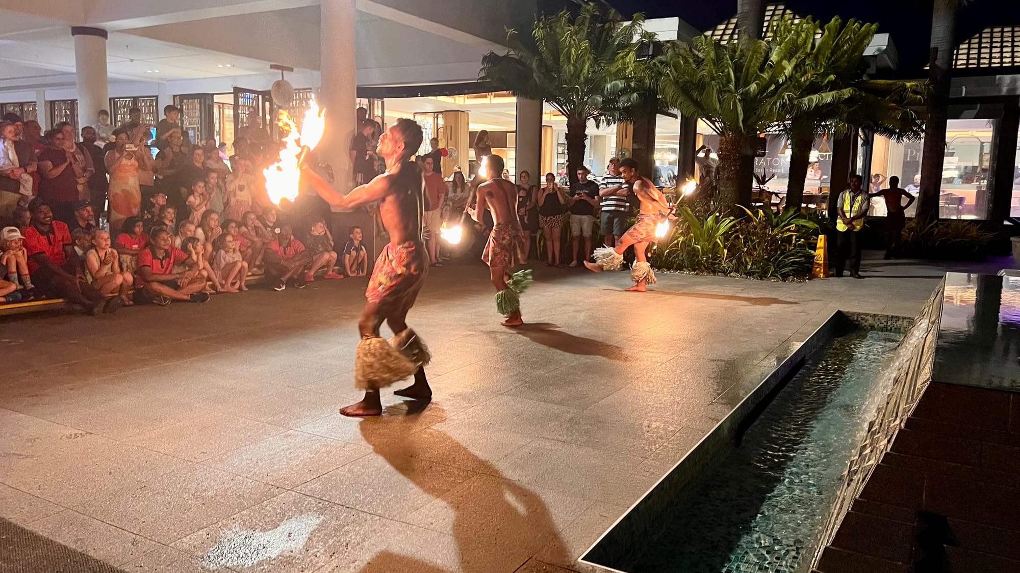Fire dancers in grass skirts spinning flaming batons on a stone patio at night, watched by a crowd seated around the edges.