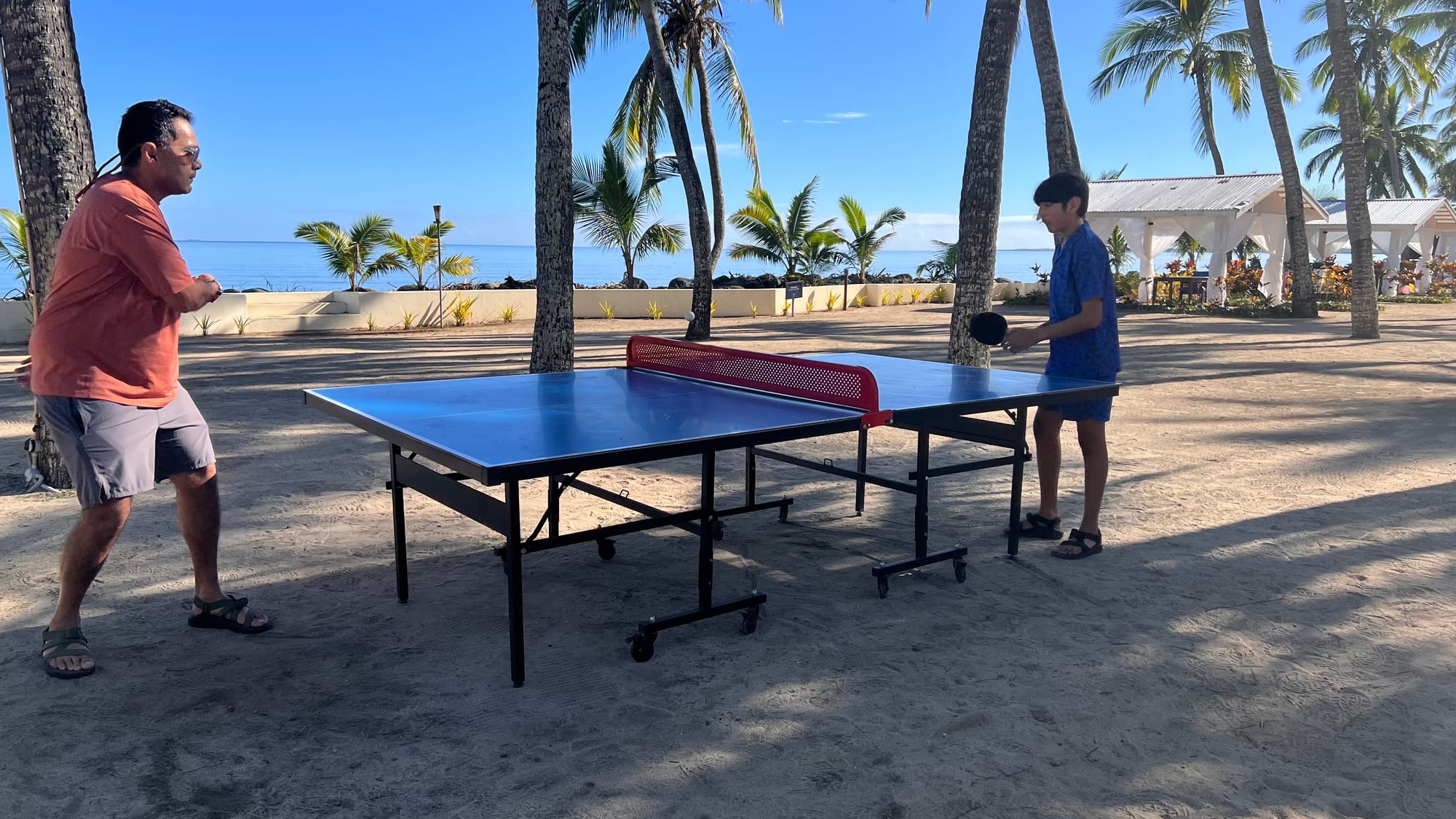 Our son and his dad playing table tennis on a blue table set up on the sand, under tall palm trees with the sea beyond.