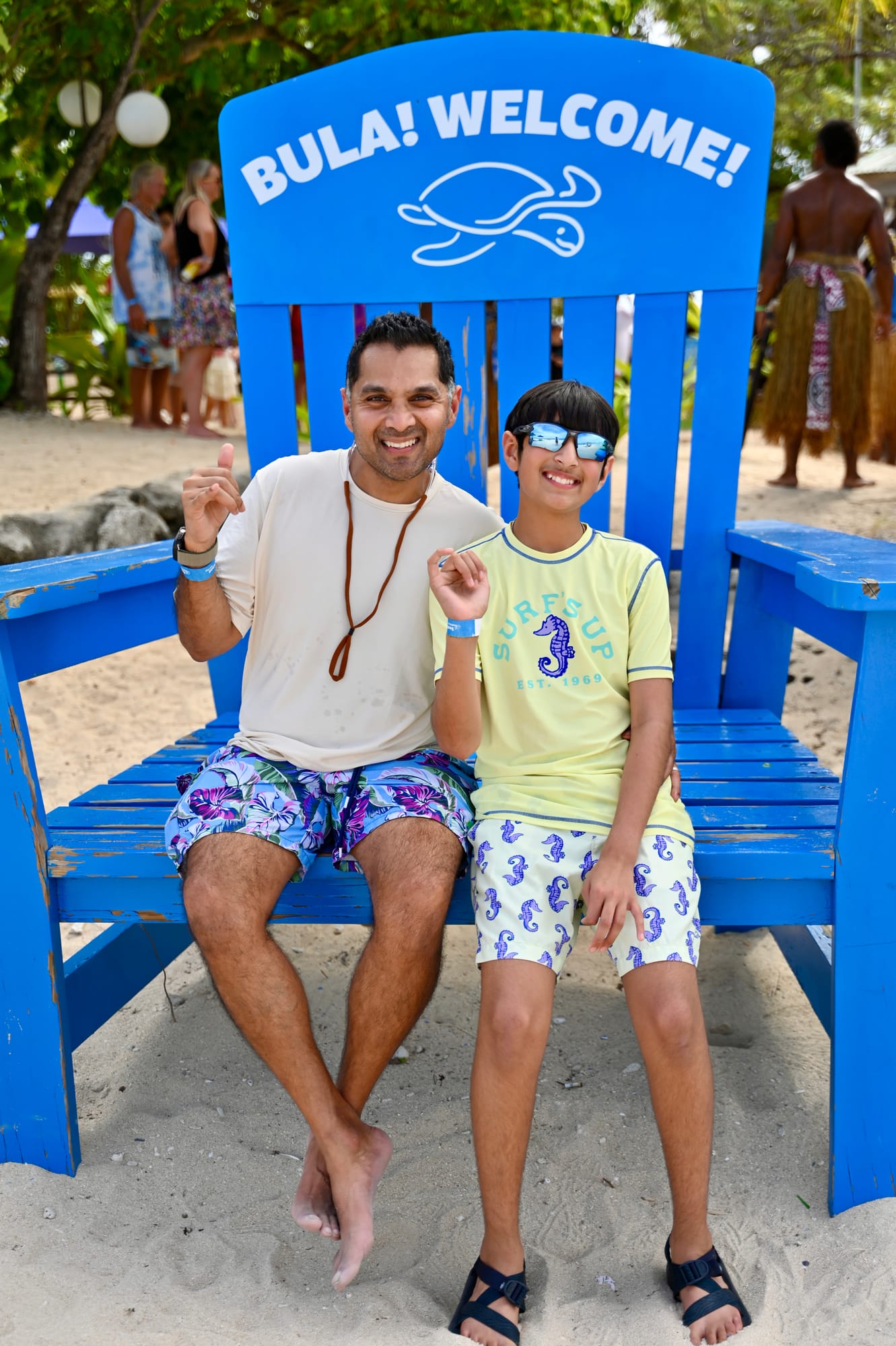 Our son and his dad sitting together on the giant blue “Bula! Welcome!” chair on the sand, grinning and throwing hand signs.