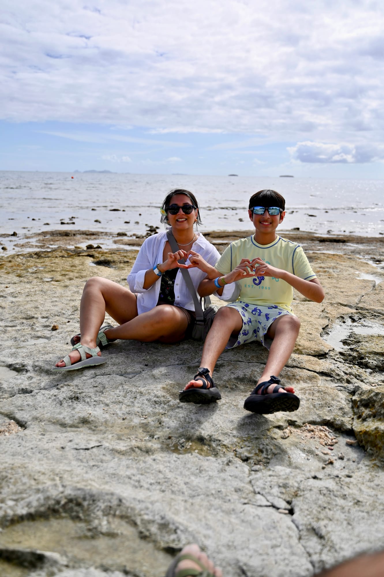 Our son and I sitting on a flat, rocky shelf at the water’s edge, making heart shapes with our hands, with the open sea and distant islands behind.