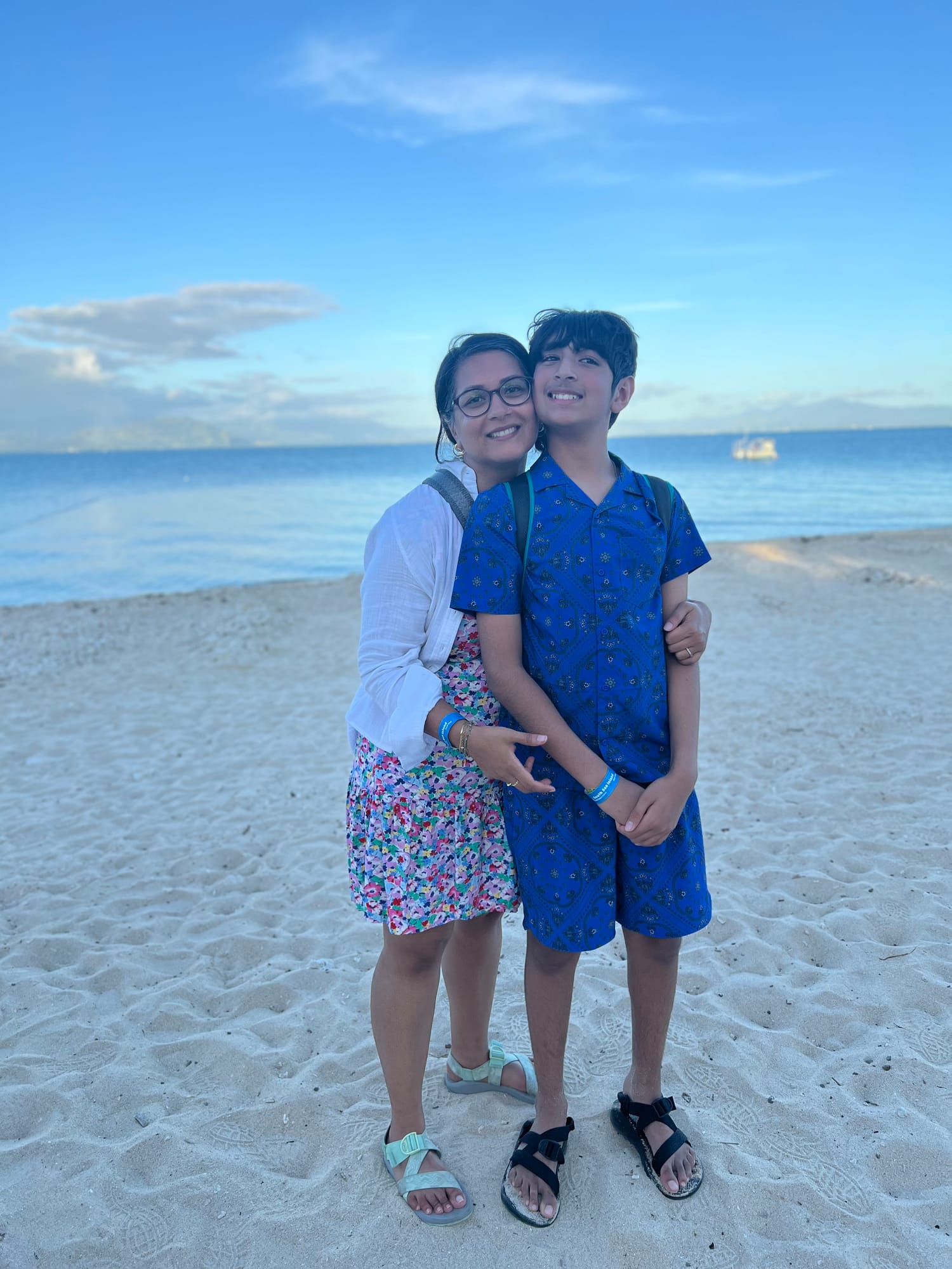 Our son and I hugging on soft white sand at the water’s edge, with calm blue sea and a pale evening sky behind.