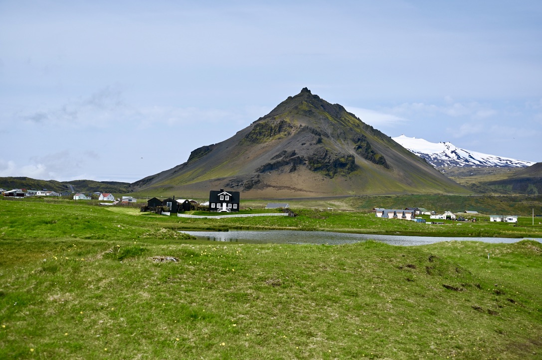 Rocky pointy mountain with snowcapped mountains in background; moss and grass covered grasslands in the front and a lake dotted by houses around it