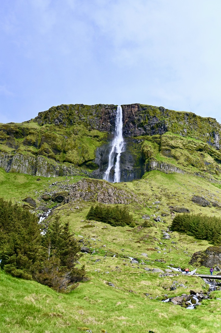 Tall cascading waterfall flows from an emerald green cliff and has basalt columns along the bottom