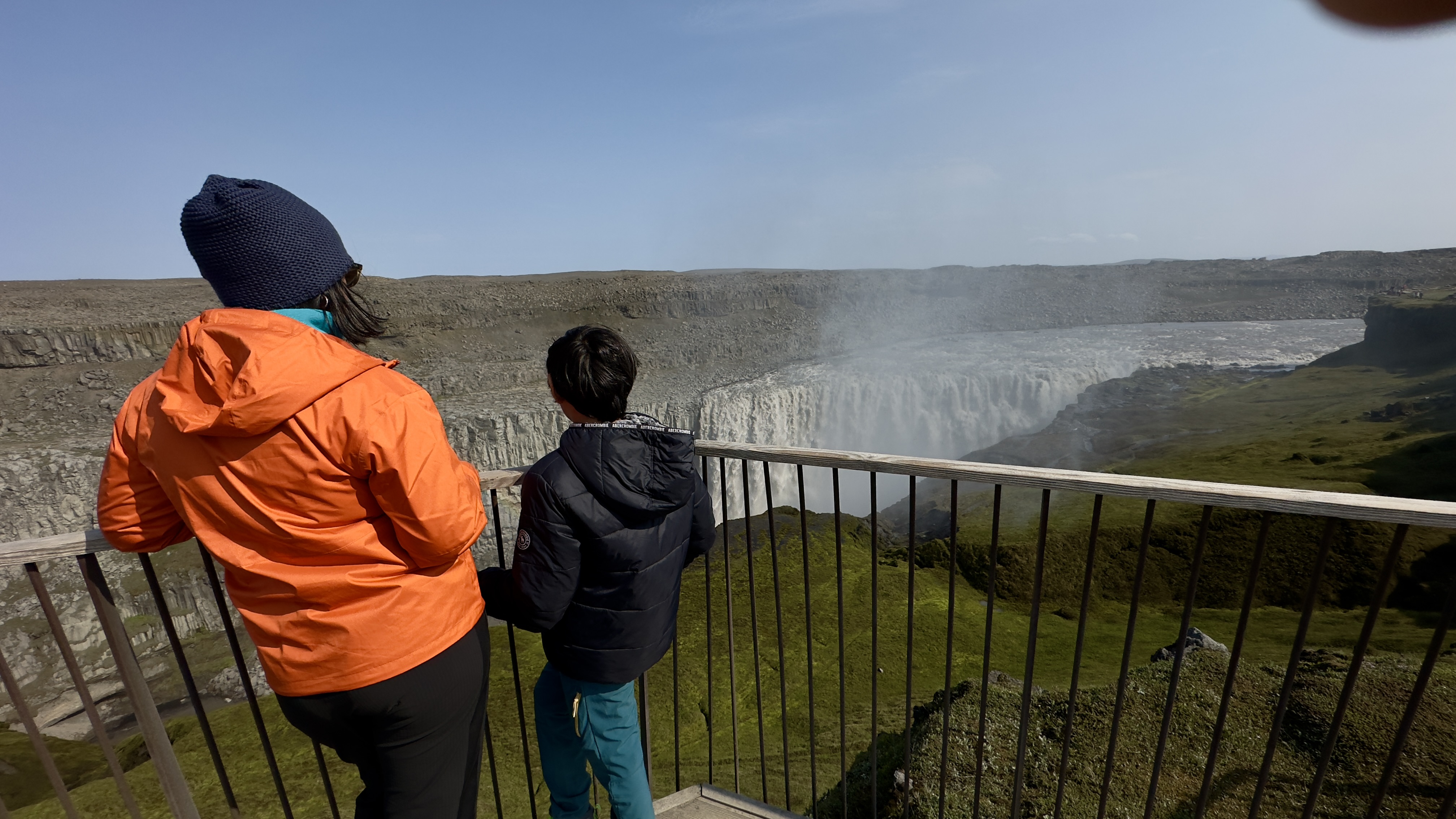 A woman and boy kid behind the railing of the waterfall named Dettifoss and the hillside is covered with moss; the other side of the waterfall is rocky and barren.
