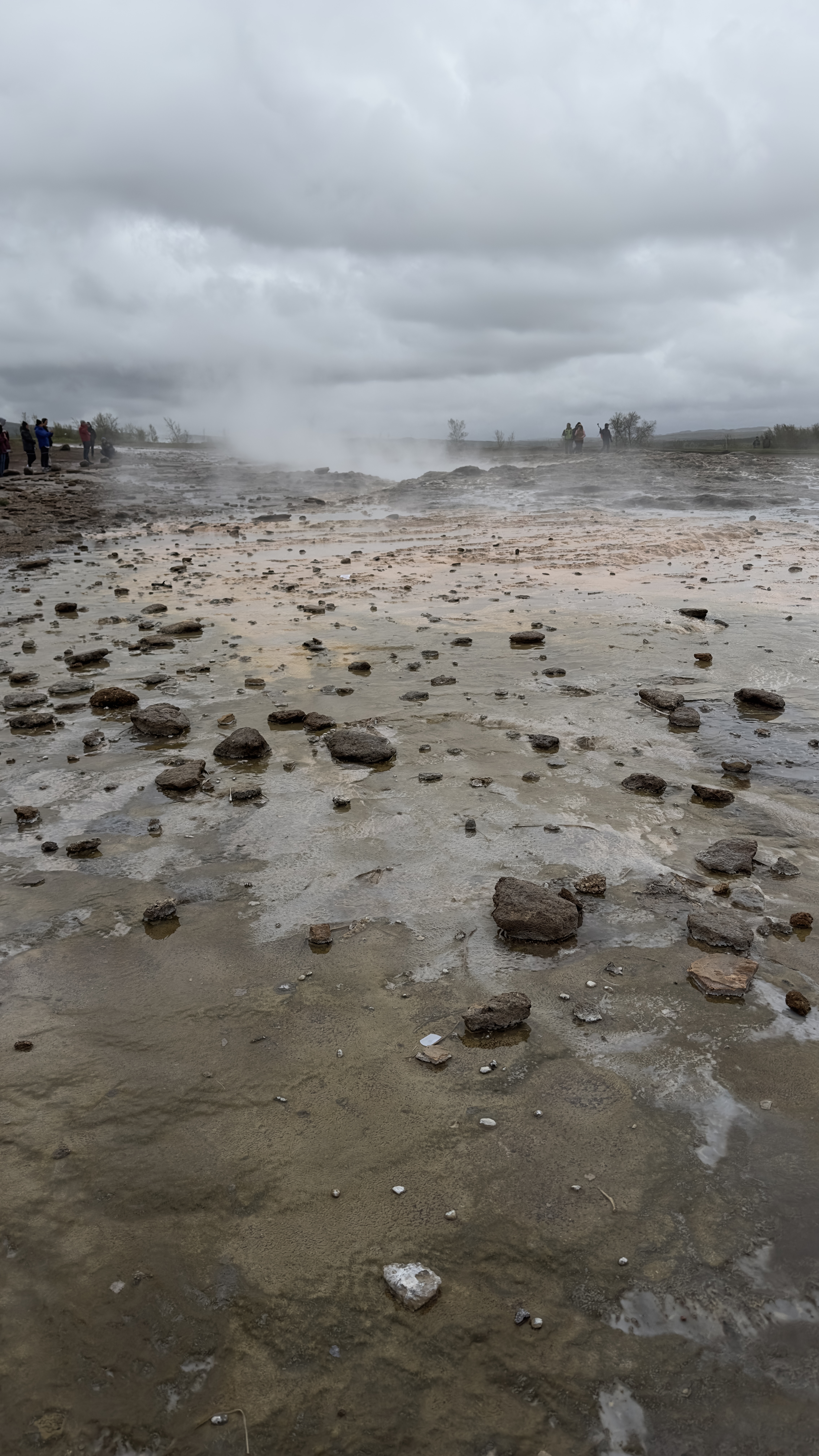 Geyser fumes coming out of the ground at the horizon with cloudy skies; wet mud and rocks strewn around the place; some people at the distance around the geyser