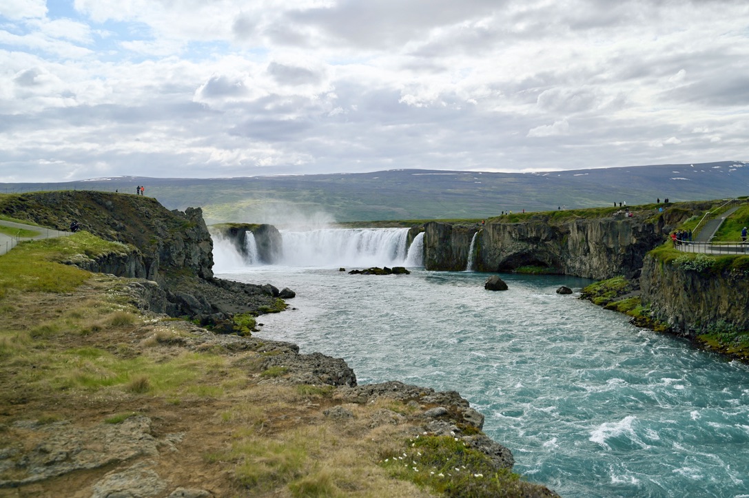 A curved wall of waterfalls with and landscape covered by moss, with mountains with snowy patches in the background
