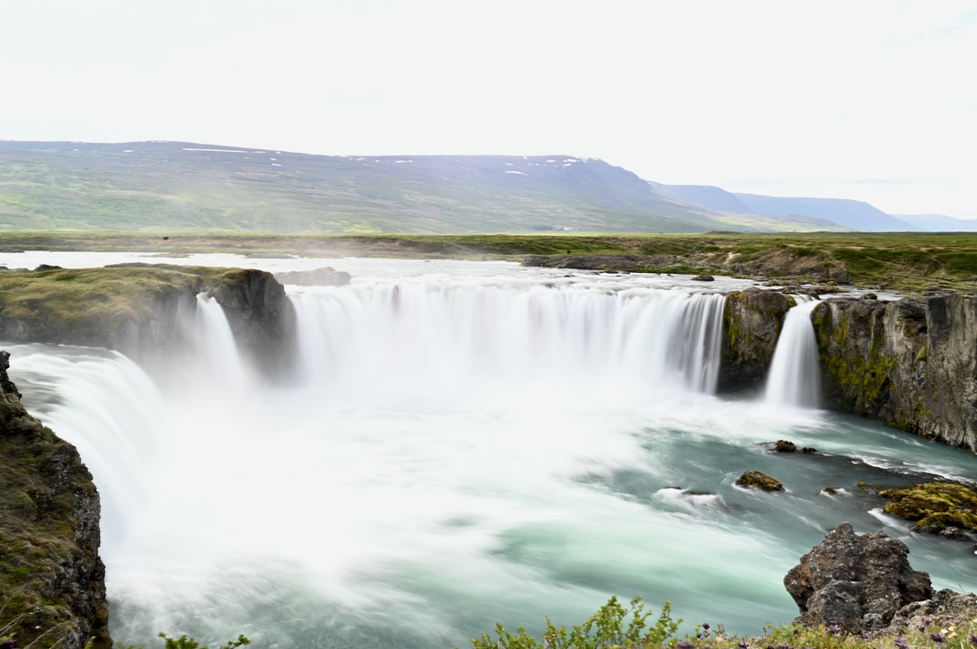 A curved wall of waterfalls with and landscape covered by moss, with mountains with snowy patches in the background