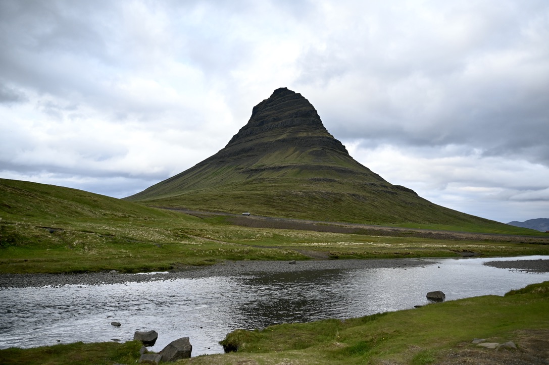 Kirkjufell mountain with a river in front. The mountain is moss and grass covered in summertime on a cloudy day.