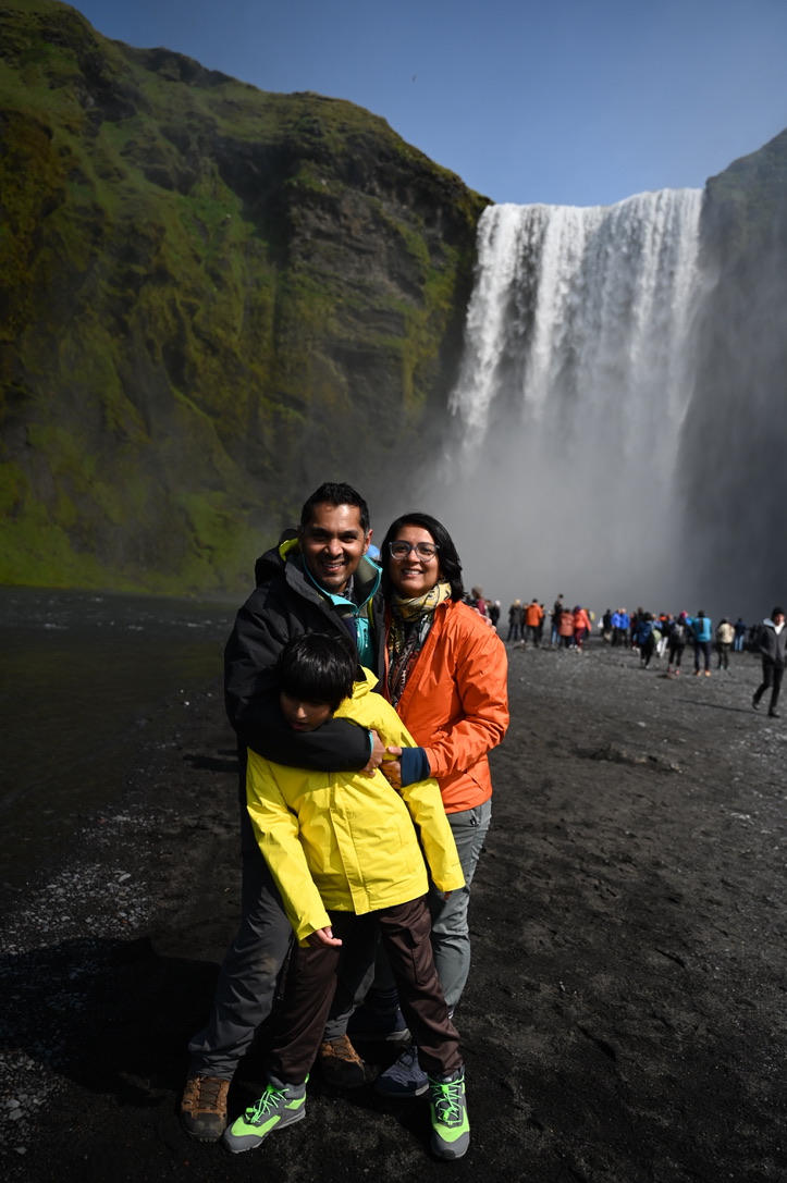 Man, woman and 10 year old boy in front of the Skogafoss waterfall in Iceland; it's coming down from a flat cliff line covered by moss and the waterfall is wide.