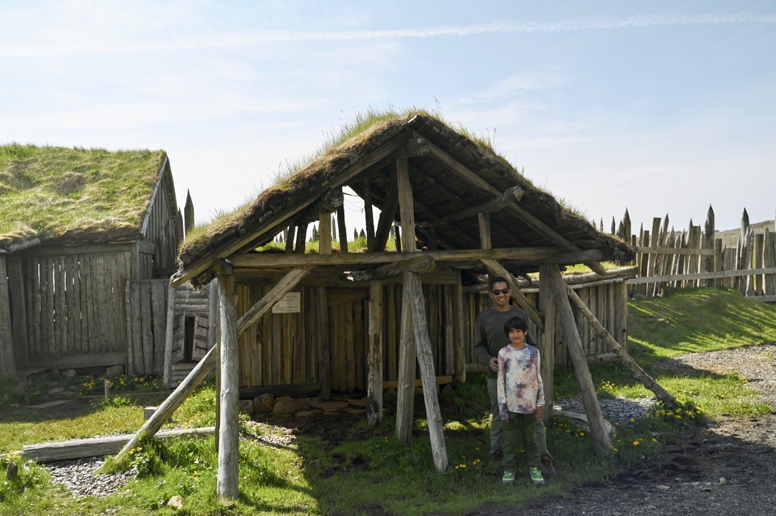 Man and kid in front of a log shelter and village with turf covering for roof. It is in the Viking village of Stokksnes