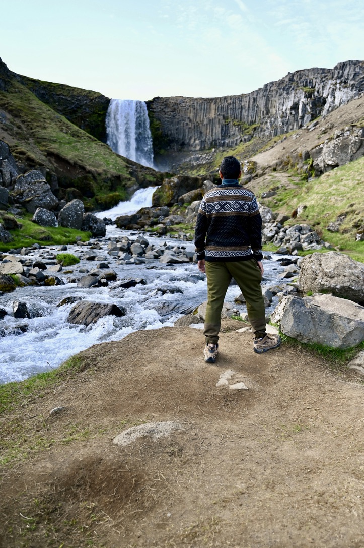 A man standing with his back facing looking at Svodufoss waterfall and the river flowing by him