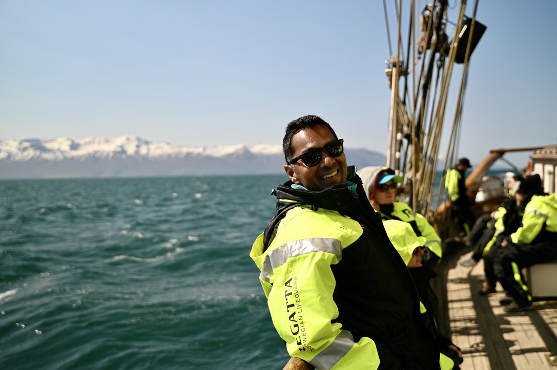 Man leaning on wooden railing of sail ship with cold ocean and snow covered mountain range in the back