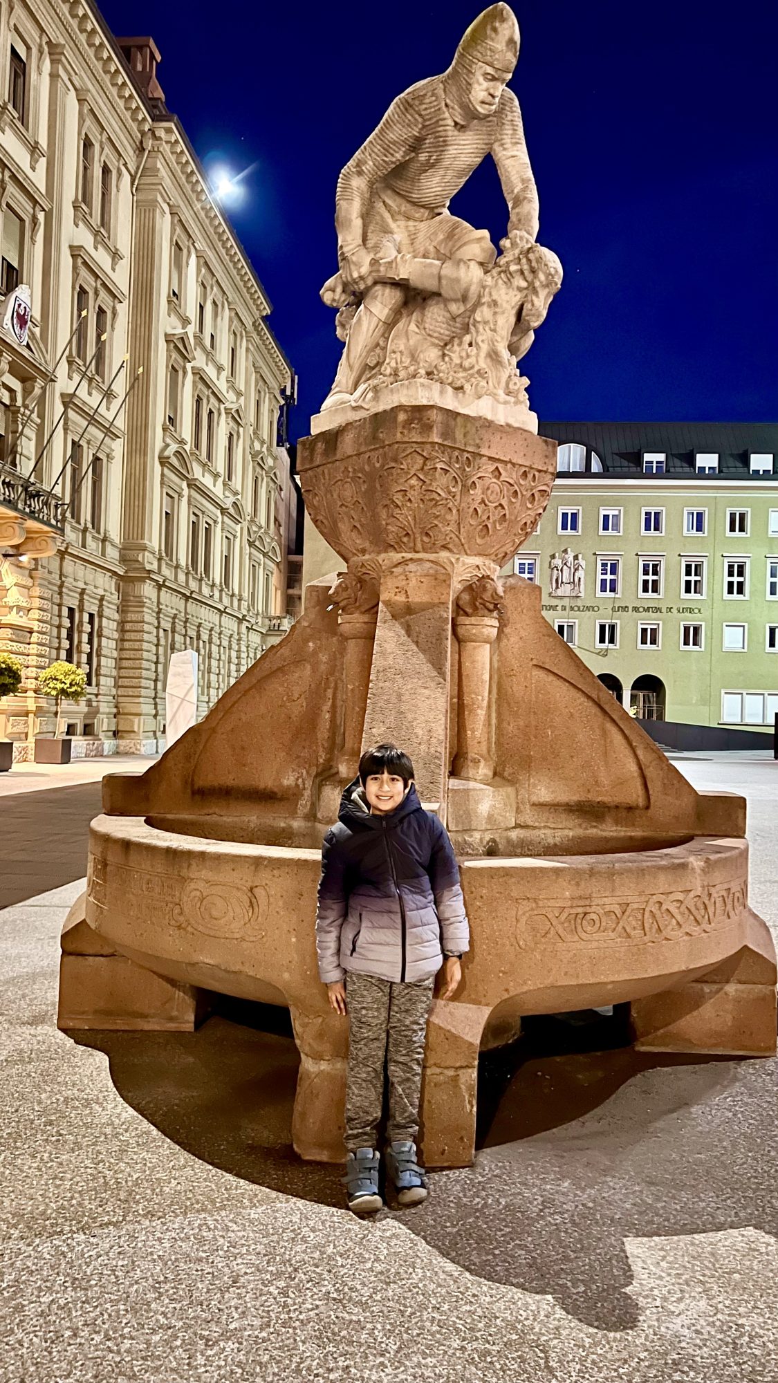 Boy standing by a carved stone fountain in Bolzano’s old town at night
