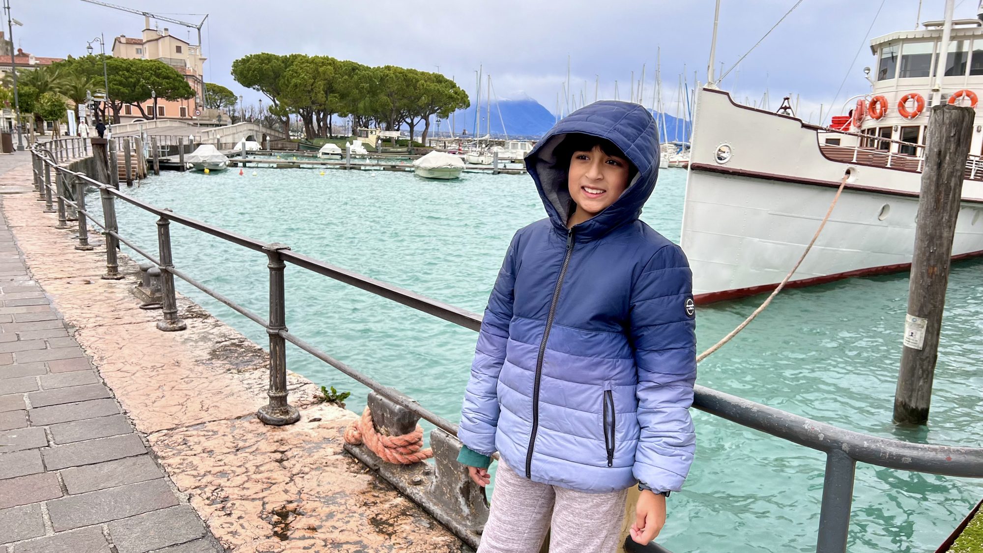 Boy in a hooded coat by the railing of Desenzano’s harbor, with boats behind