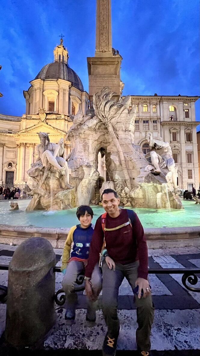 Man and boy sitting in front of the lit-up Fountain of the Four Rivers at dusk