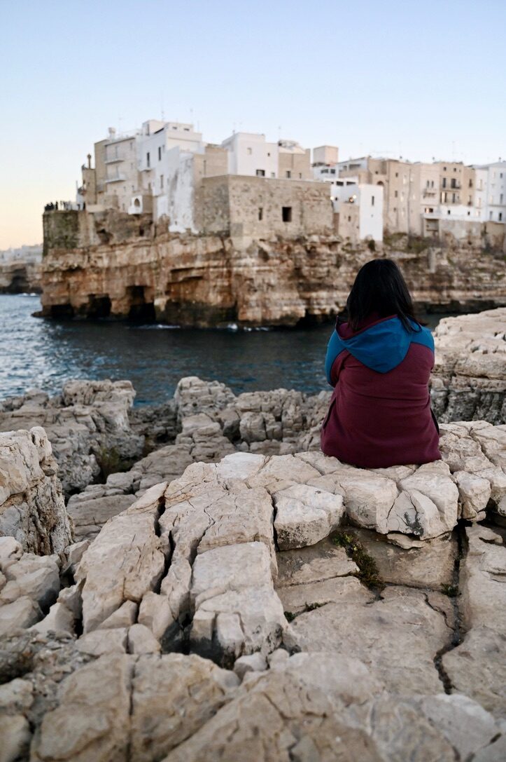 Woman sitting on the rocks facing the white town on the cliffs of Polignano a Mare