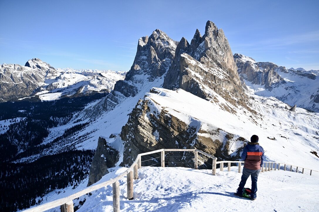 A person at a railing looking at the sharp, snow-dusted Seceda ridge