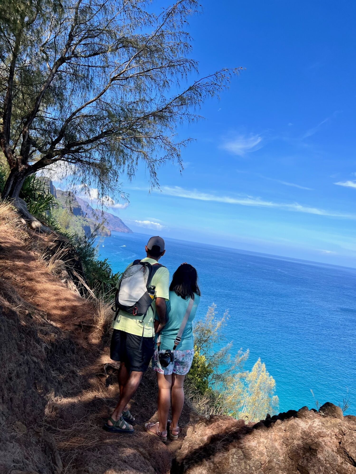 Man and woman standing at the edge of the Kalalau Trail, looking out over the blue Pacific and the Napali cliffs