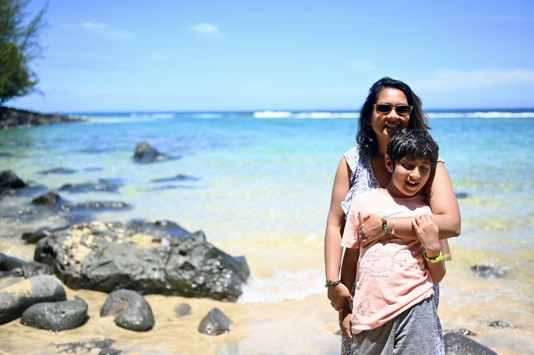 Woman hugging a boy on the sand at Keʻe Beach, with dark rocks and clear turquoise water behind them