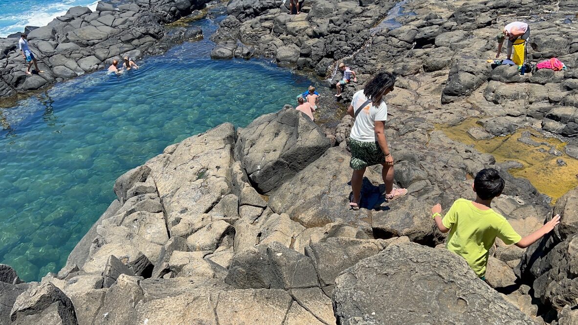 Woman and boy climbing down black lava rocks to the clear green pool of the Queen’s Bath