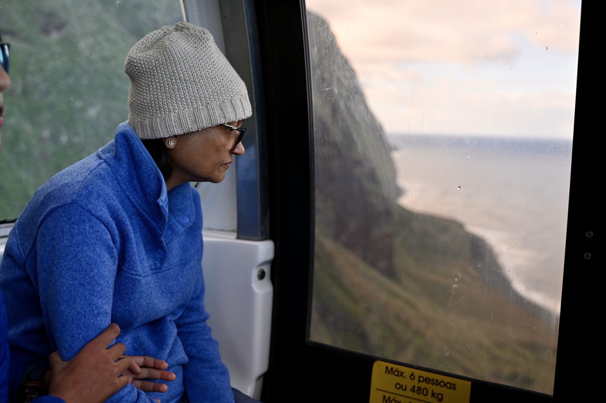 One of us in a grey beanie looking out of the cable car window at the cliff face and the sea far below.