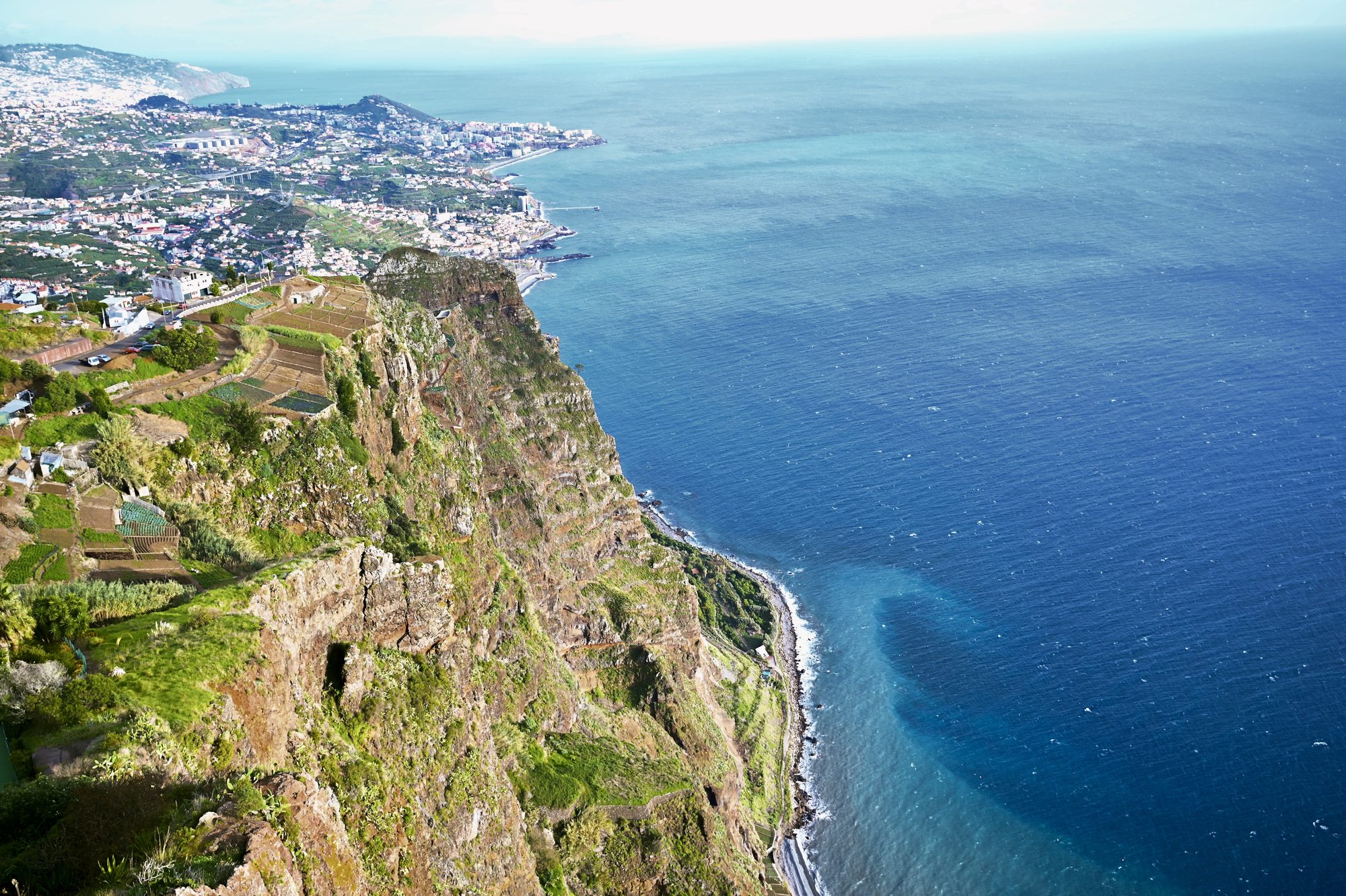 Looking straight down a sheer sea cliff to terraced fields and the deep blue ocean, with Funchal spread along the coast.