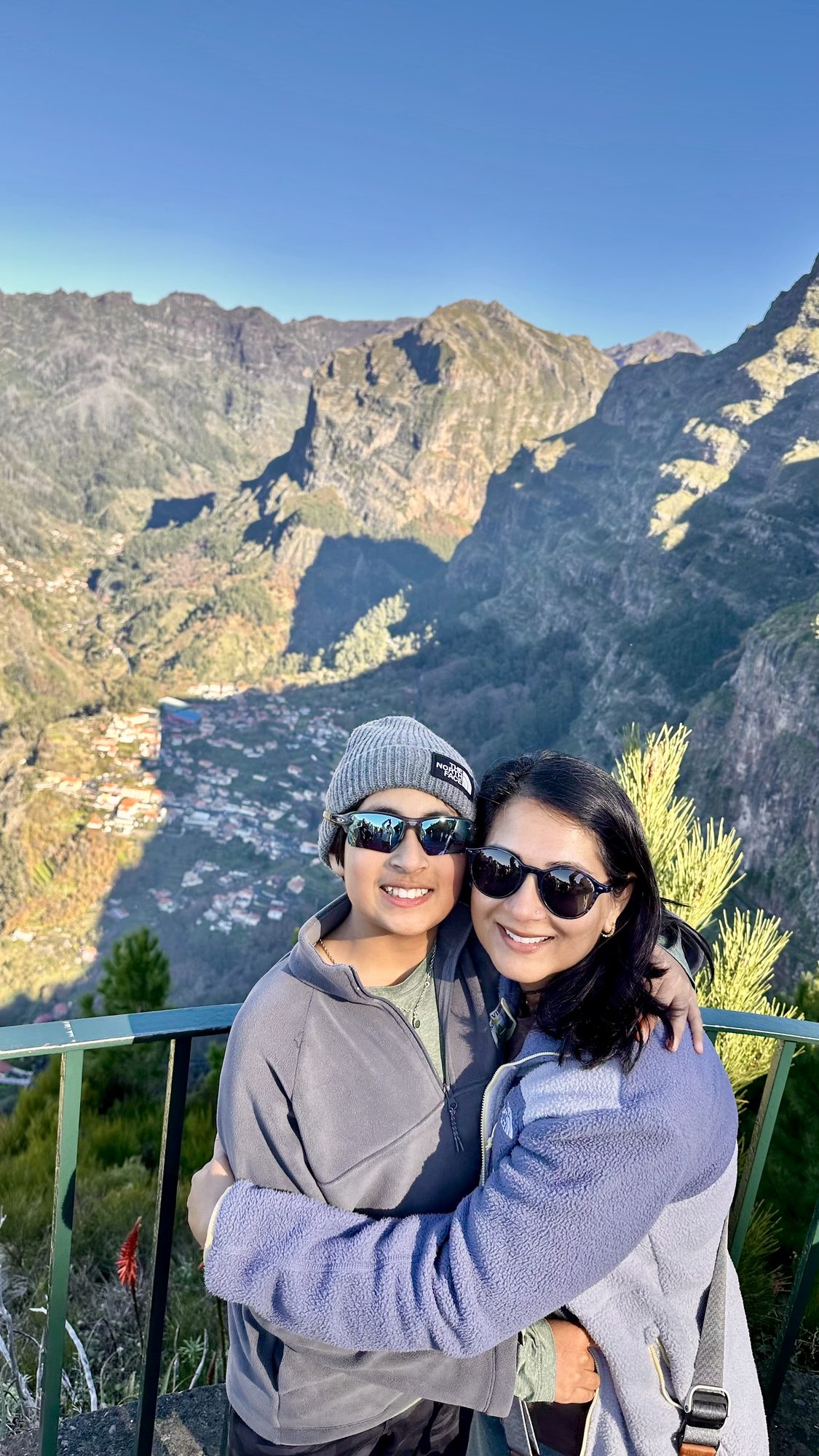 Two of us hugging at the railing, with sharp mountain ridges and the valley far below.