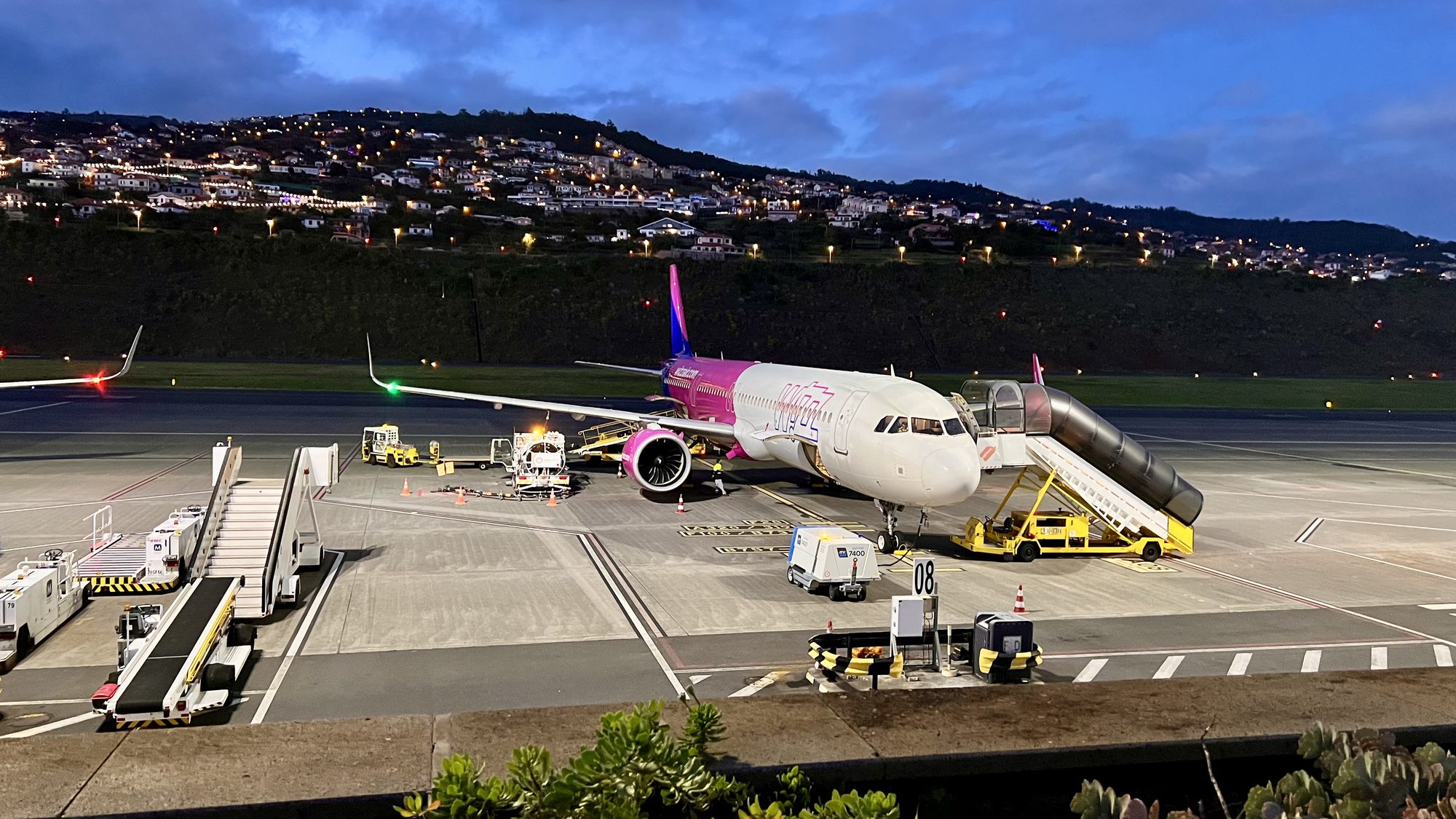 A plane parked on the apron at dusk, with the lights of the hillside town behind.